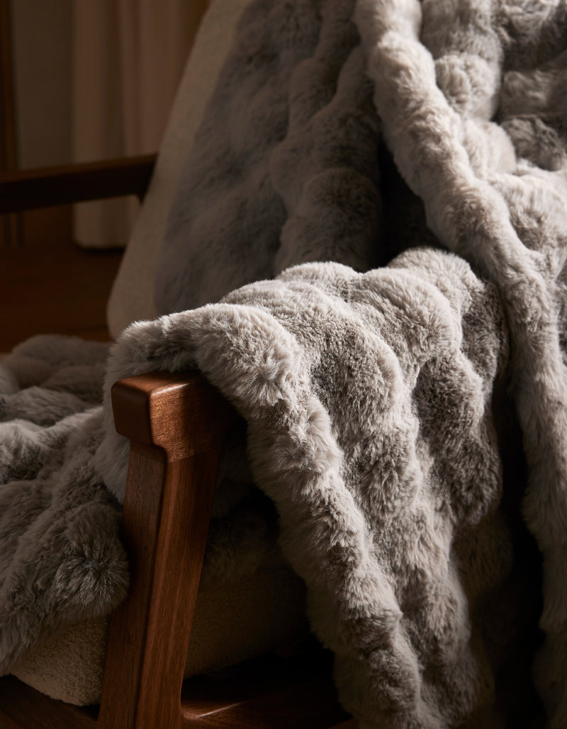 The Bubble Cuddle Blanket by Cozy Earth, with its soft gray faux fur, is draped over a wooden chair, adding a cozy and inviting touch to the space.