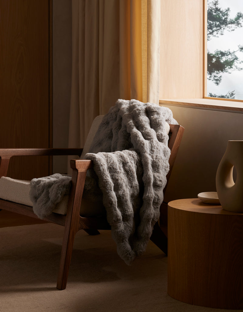 A wooden armchair with a beige cushion is draped with the Cozy Earth Bubble Cuddle Blanket in textured gray. Beside it, a round wooden side table holds a sculptural beige vase, while natural light from the window reveals a tree outside.
