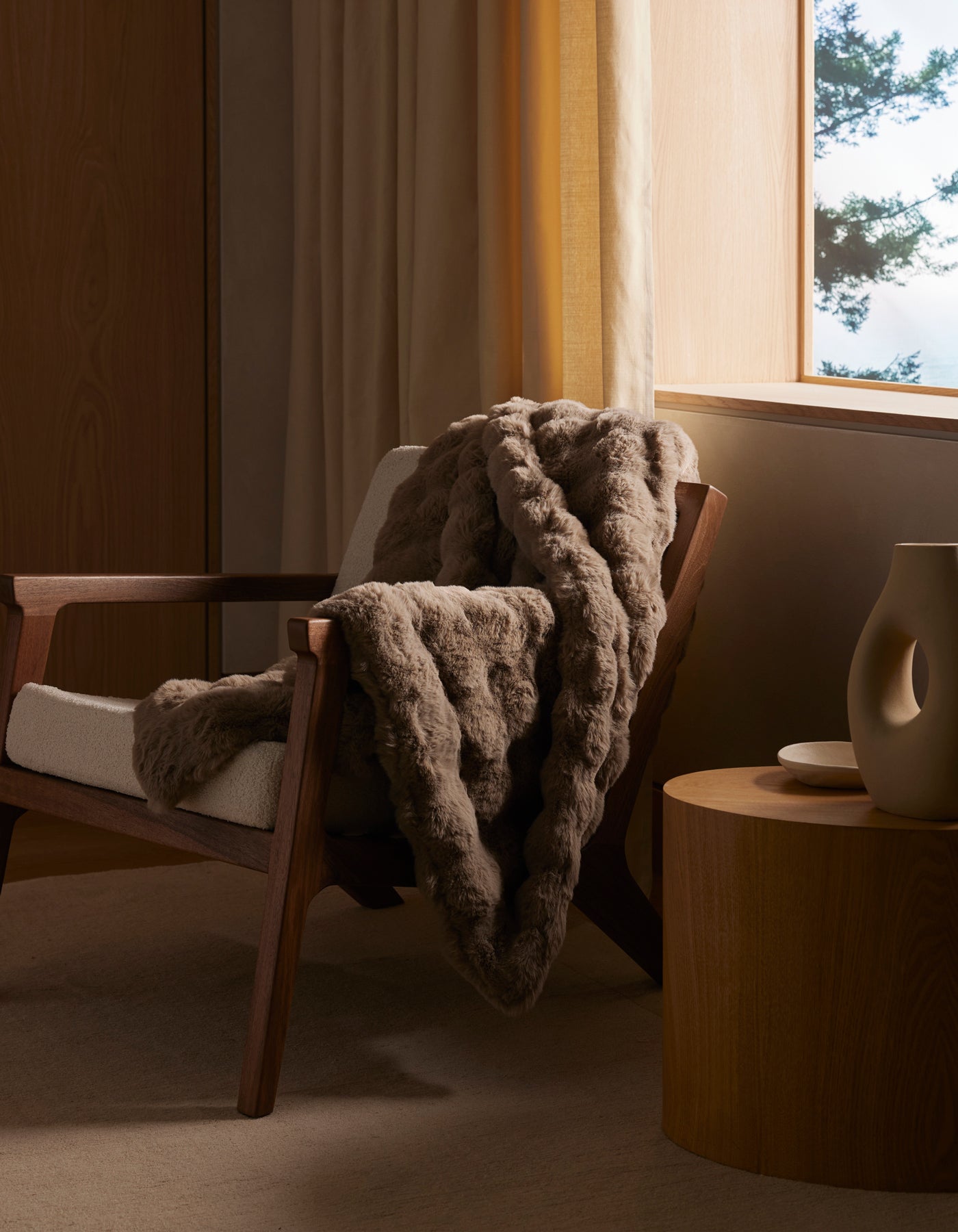 A cozy wooden armchair with a cream cushion and a Bubble Cuddle Blanket by Cozy Earth draped over it sits next to a round wooden side table with minimalist ceramic decor, beside a sunlit window with beige curtains.|Color:Walnut