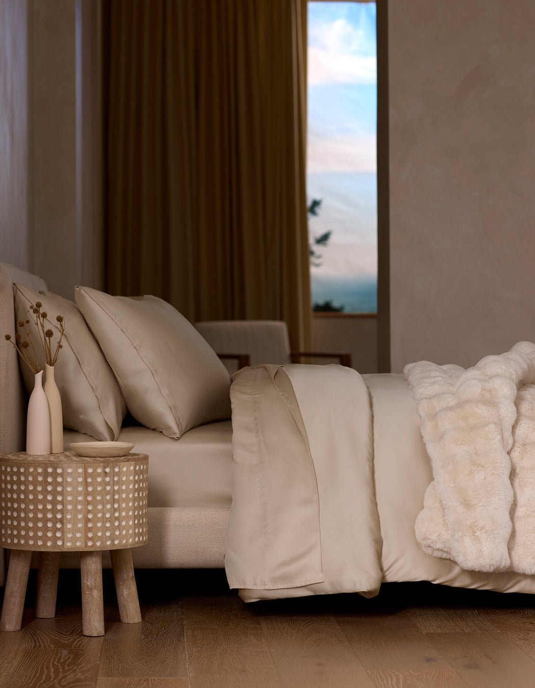 A neatly made bed features the Cozy Earth Bamboo Duvet Cover in beige and a white faux fur blanket, beside a textured wooden nightstand with two vases. Large curtained windows reveal a scenic sky.