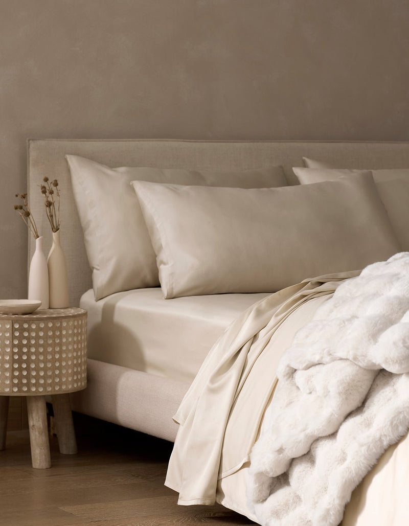A neatly made bed features Cozy Earth Bamboo Pillowcases in cream, a soft white blanket, and a nightstand with minimal vases of dried plants against a beige wall.