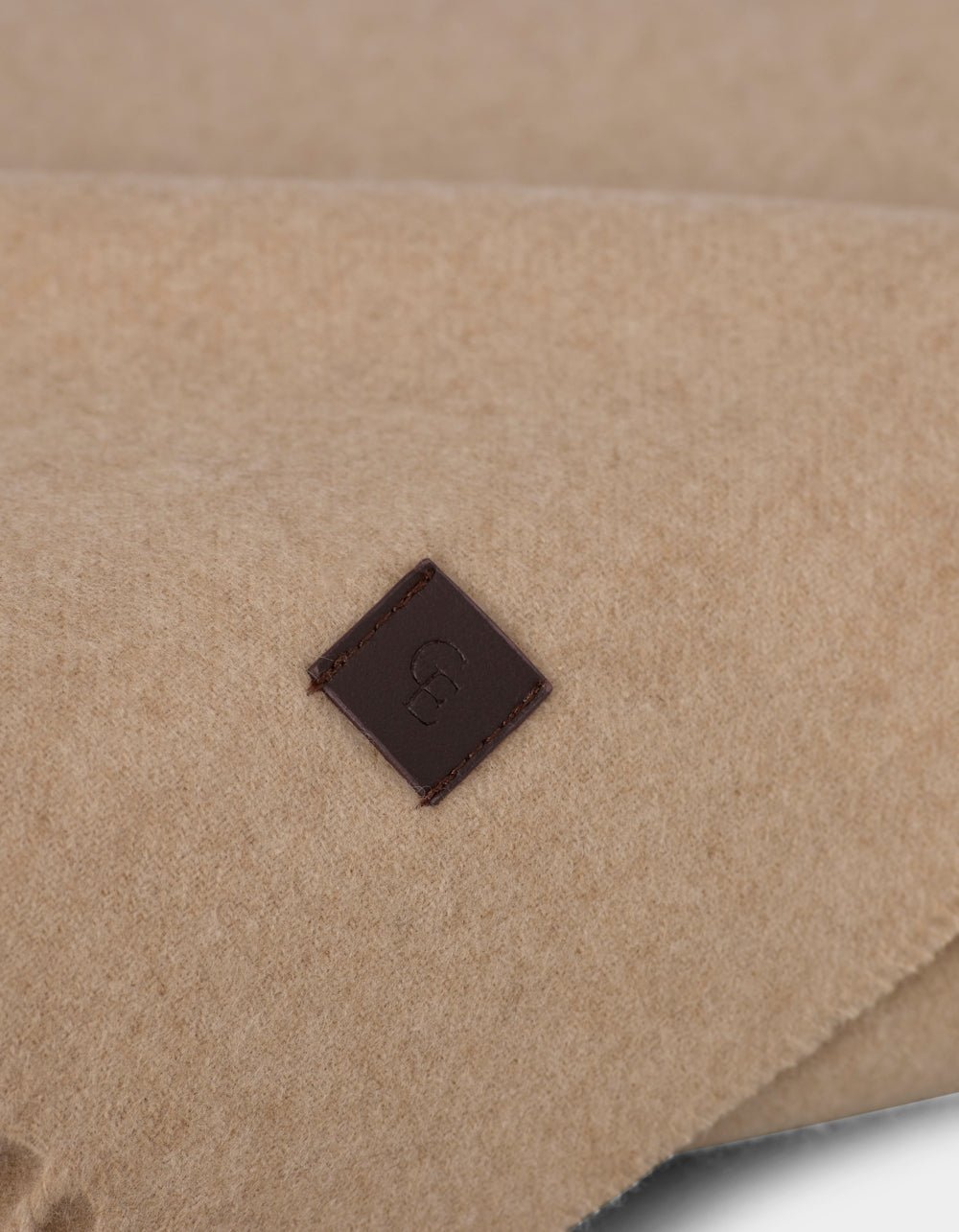 Close-up of the Cashmere Tassel Throw by Cozy Earth, featuring soft beige fabric and a small square brown leather tag stitched on, embossed with a simple logo.