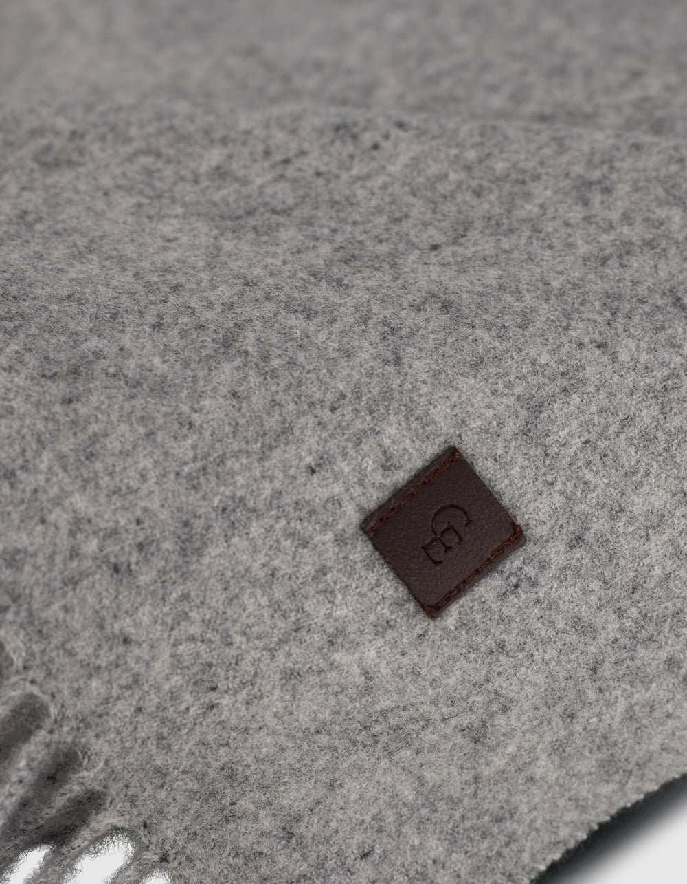 Close-up of Cozy Earth’s Cashmere Tassel Throw in soft light grey, showing its cozy texture. A small brown leather tag with an embossed logo is stitched on for a refined touch.