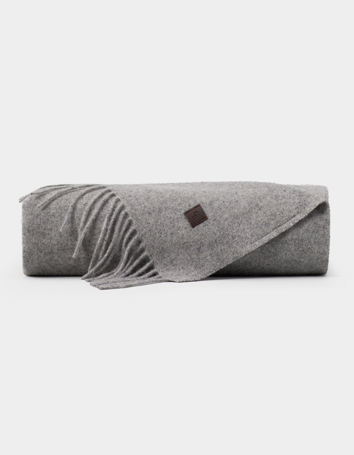 The Cozy Earth Cashmere Tassel Throw, a light gray wool blanket with fringed edges and a small brown leather patch, is neatly rolled and displayed on a plain white background.|Color:Pebble
