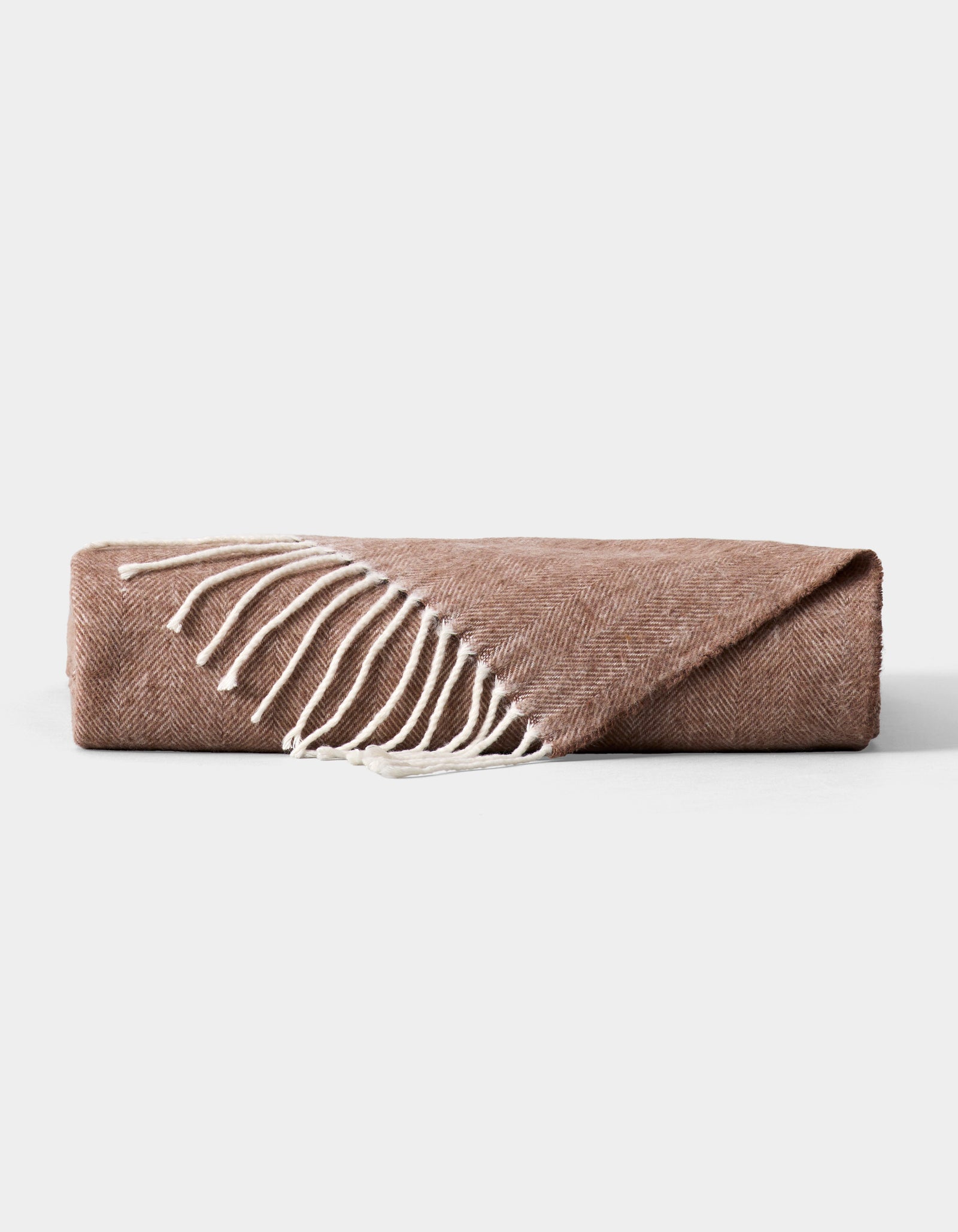 The Cozy Earth Bamboo Herringbone Tassel Throw, in light brown with a subtle herringbone pattern and white fringe, is shown folded on a plain gray background.