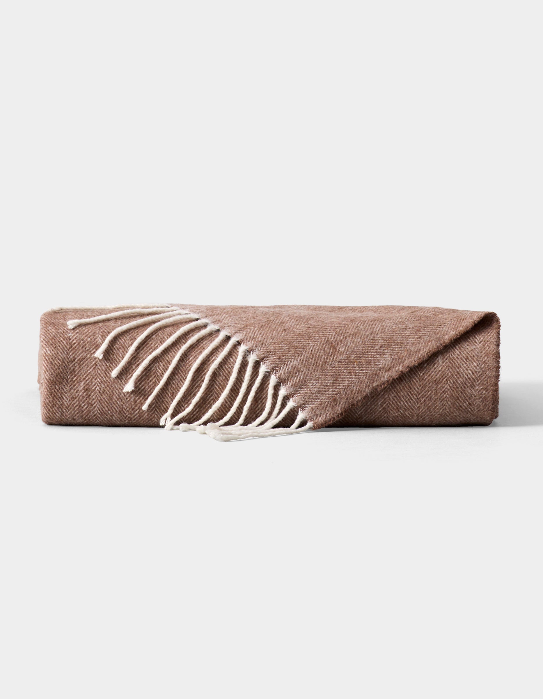 The Cozy Earth Bamboo Herringbone Tassel Throw, in light brown with a subtle herringbone pattern and white fringe, is shown folded on a plain gray background.|Color:Truffle