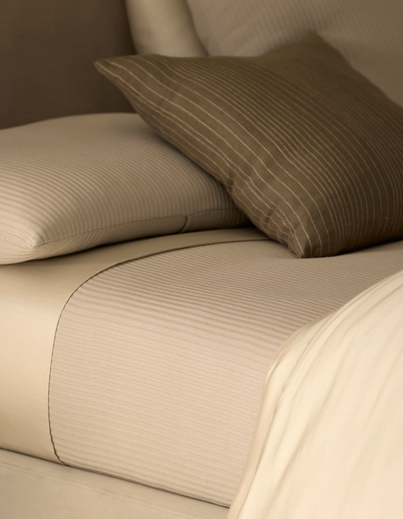 Close-up of a neatly made bed featuring the Cozy Earth Bamboo Coverlet in cream with subtle stripes, paired with a matching pillow and a brown accent pillow adorned with white pinstripes.