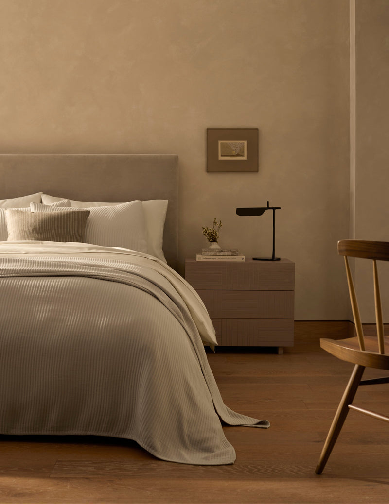 A minimalist bedroom features a neatly made bed with the Cozy Earth Bamboo Coverlet in beige tones, a wooden nightstand topped with a modern black lamp and small vase, a framed picture on the wall, and a wooden chair on hardwood floors.