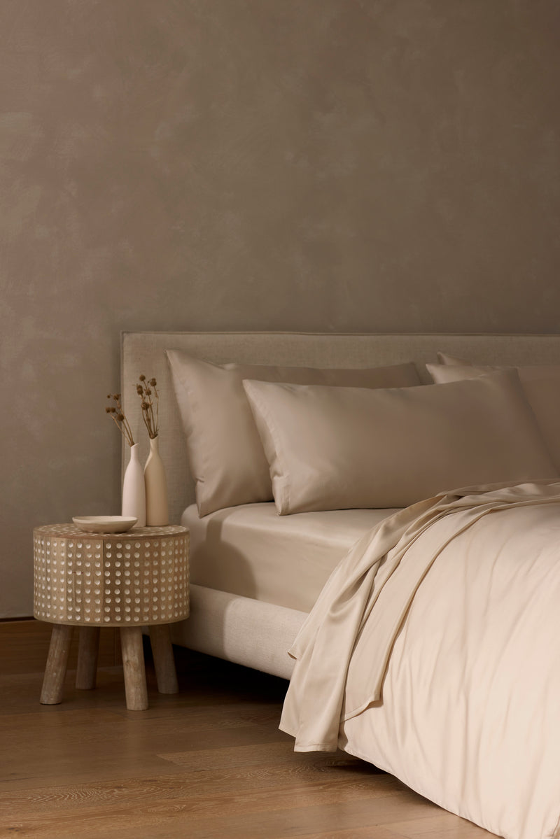 Minimalist bedroom featuring the Cozy Earth Bamboo Bedding Core Bundle on a beige upholstered bed, with a round wooden nightstand holding two white vases and a dish, set against a textured taupe wall and wooden floor.