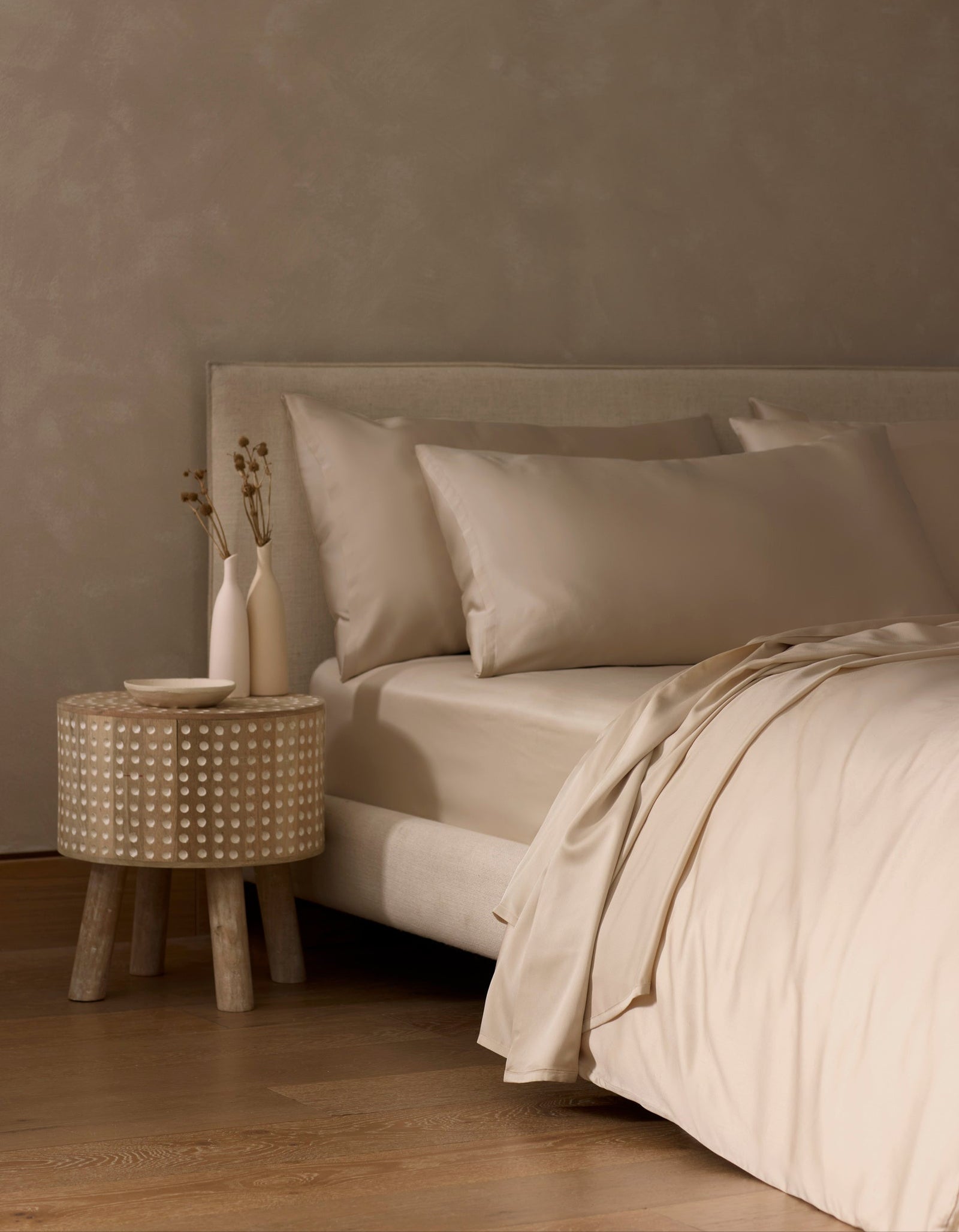 A minimal beige bedroom showcases a neatly made bed with smooth, light Bamboo Sheet Set by Cozy Earth. A small round side table with textured pattern holds two simple vases with dried flowers. The space features wooden flooring and neutral walls.