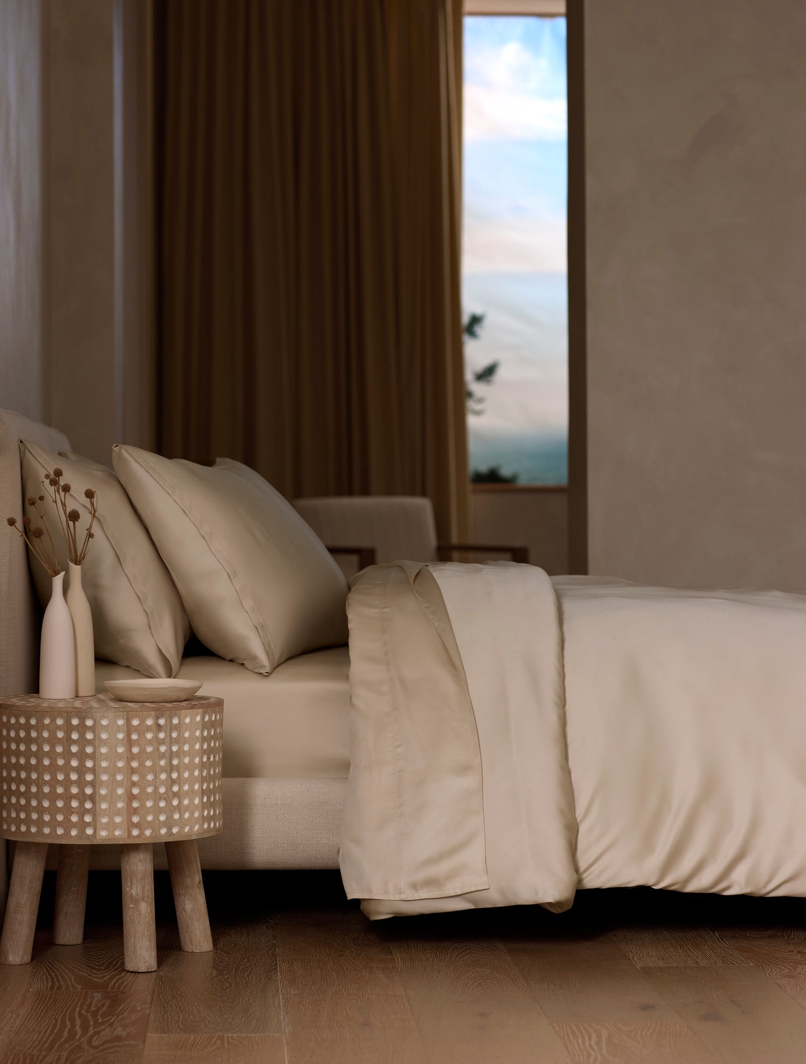A modern bedroom features a neatly made bed with Cozy Earth’s Bamboo Sheet Set in cream, a wooden side table with dried flowers, neutral walls, wooden flooring, and a window revealing a blue sky with clouds.