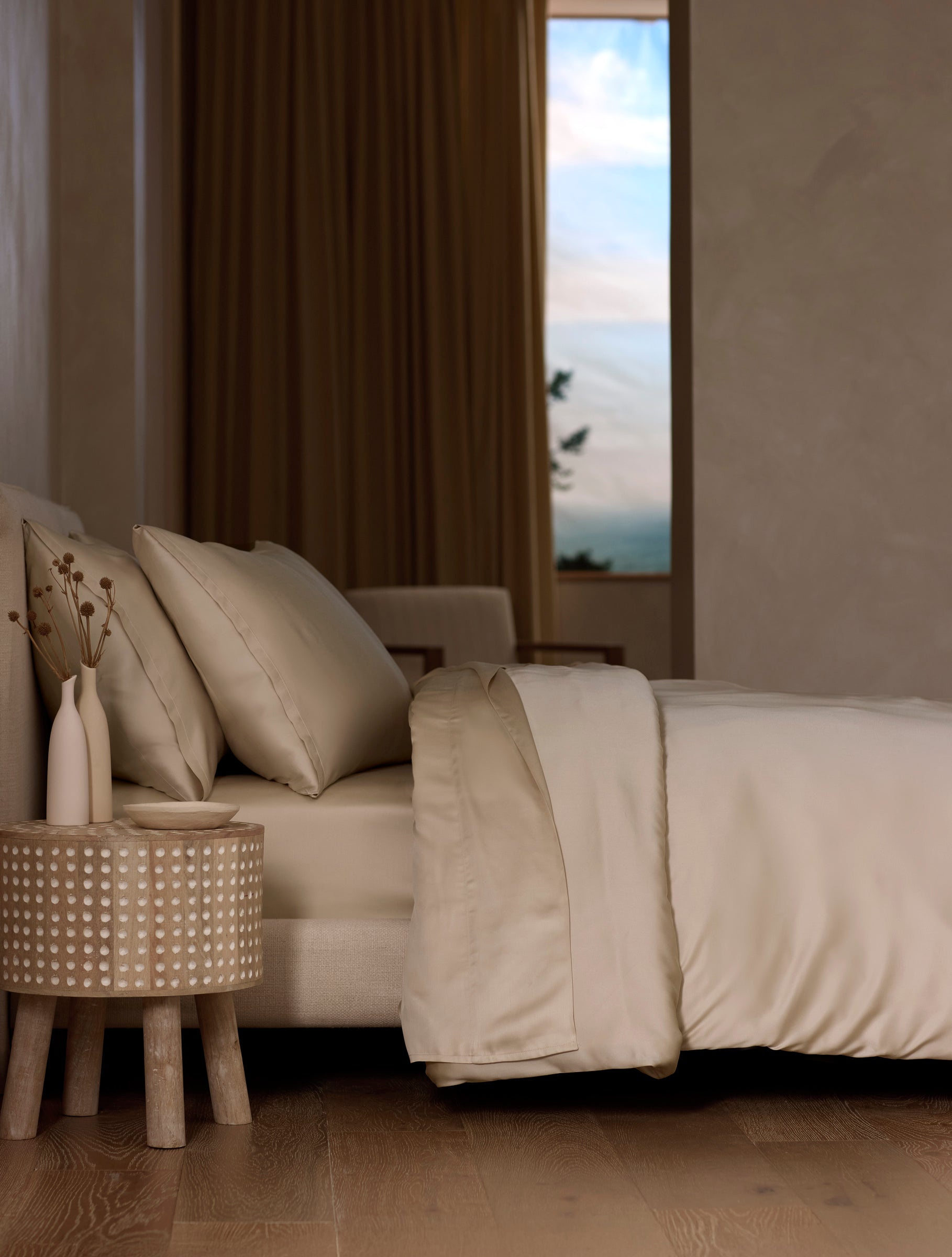 A modern bedroom features a neatly made bed with Cozy Earth’s Bamboo Sheet Set in cream, a wooden side table with dried flowers, neutral walls, wooden flooring, and a window revealing a blue sky with clouds.