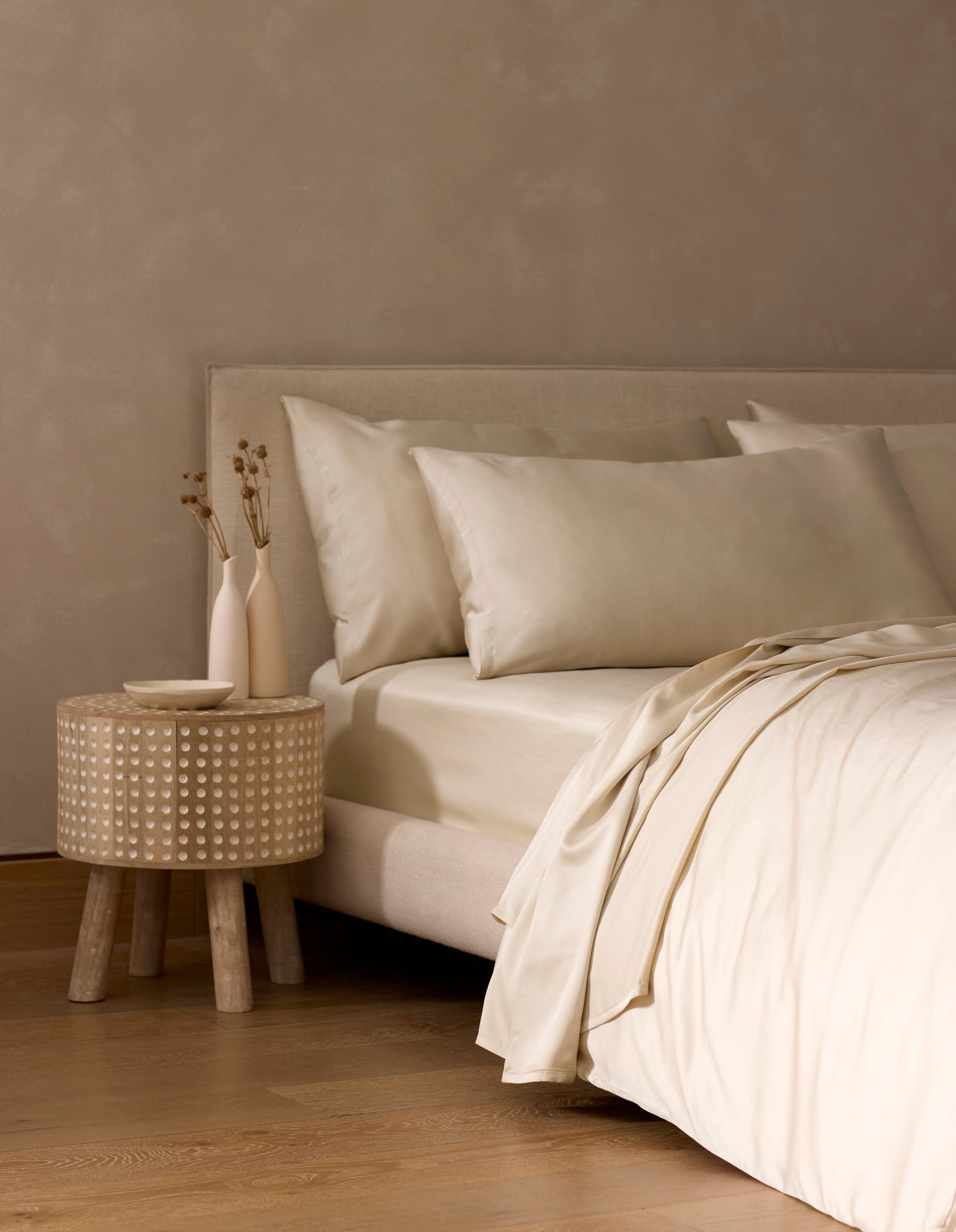 A minimalist bedroom features a beige bed with Cozy Earth's Bamboo Bedding Core Bundle in cream, a round wooden nightstand, two vases with dried flowers, and a neutral wall over a wooden floor.