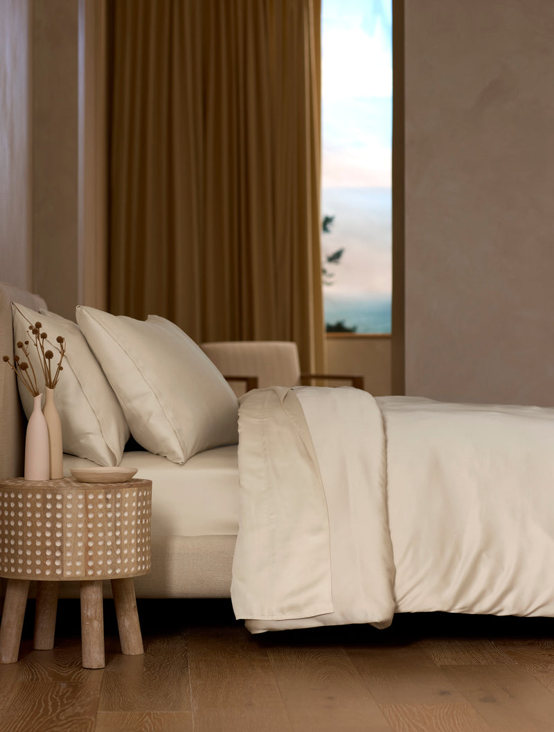 A bed dressed in Cozy Earth’s Bamboo Bedding Deluxe Bundle features cream-colored sheets and pillows, set beside a round wooden table with dried flowers. In the background are a beige armchair and a window with gold curtains.