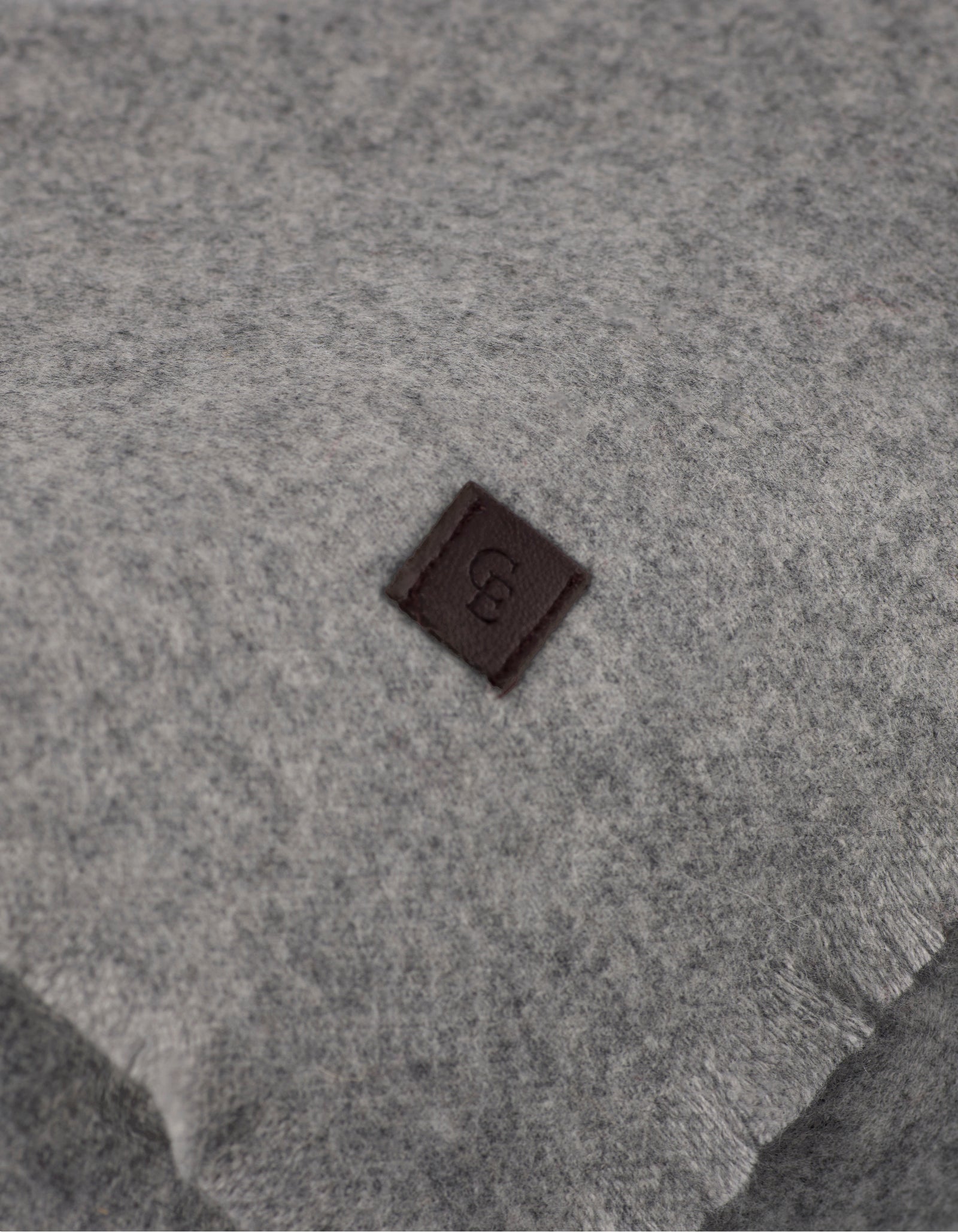 A close-up of Cozy Earth's Cashmere Fringe Blanket shows its soft, light gray fabric accented with a small, dark brown square patch embossed with a "B.