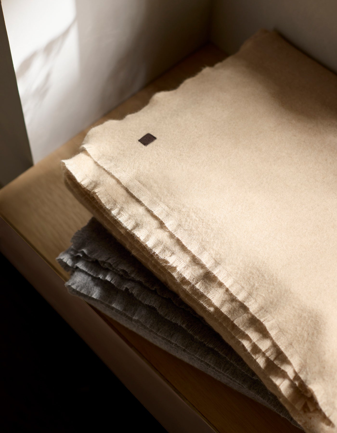 A Cozy Earth Cashmere Fringe Blanket in beige is neatly folded atop a gray one on a wooden surface, with soft light casting gentle shadows around them.