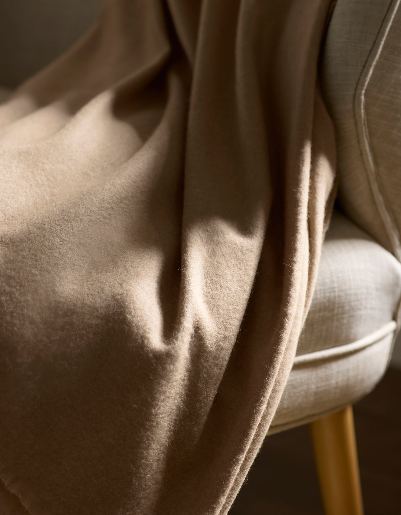 The Cashmere Fringe Blanket by Cozy Earth, in soft light brown, is draped over a light beige upholstered chair with wooden legs and bathed in natural sunlight.
