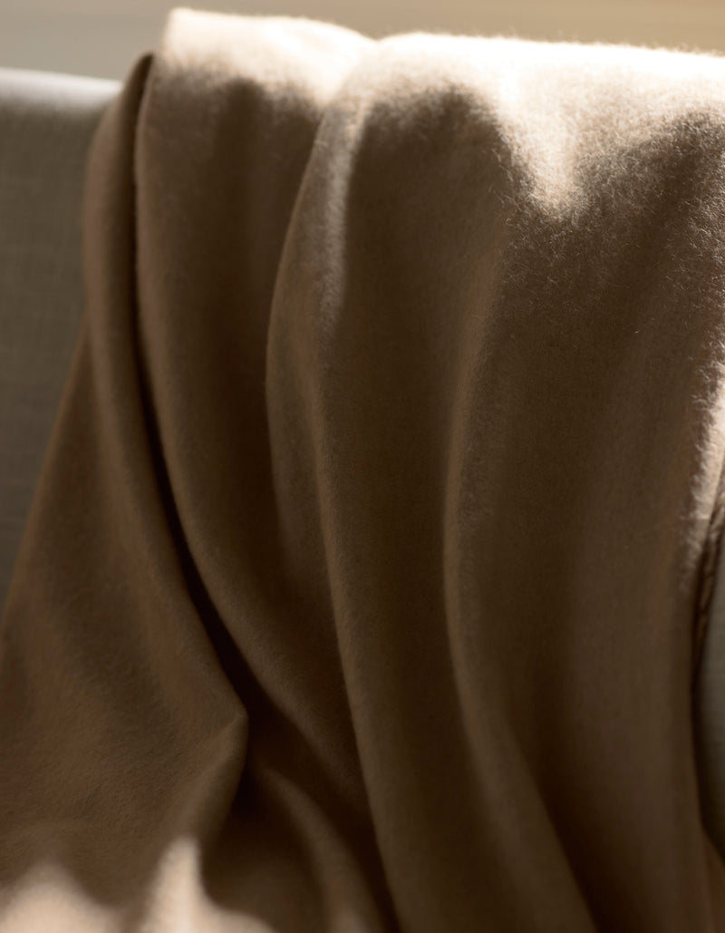 The Cozy Earth Cashmere Fringe Blanket, in a soft brown shade, is draped over a light-colored sofa as sunlight casts gentle highlights and shadows on its luxurious fabric.