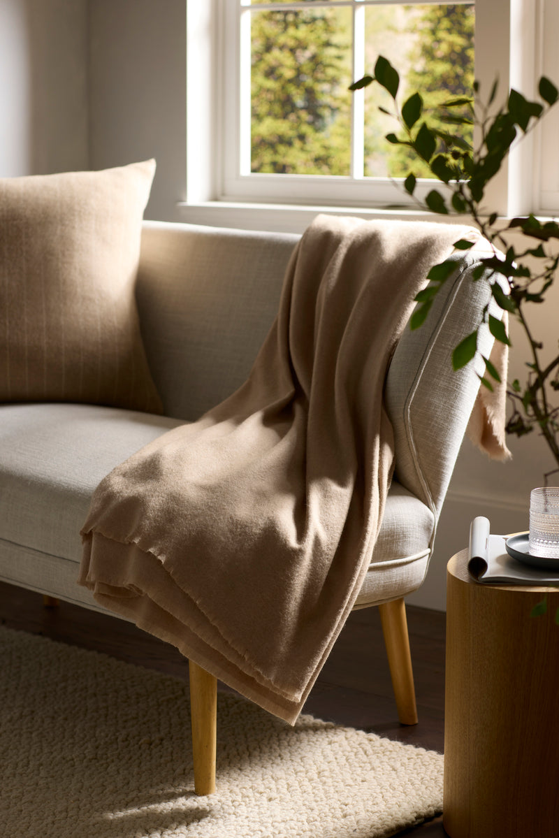 The Cozy Earth Cashmere Fringe Blanket in beige is draped over a light gray sofa with a matching cushion, beside a leafy plant and a side table with a glass of water, in a bright, sunlit living room.