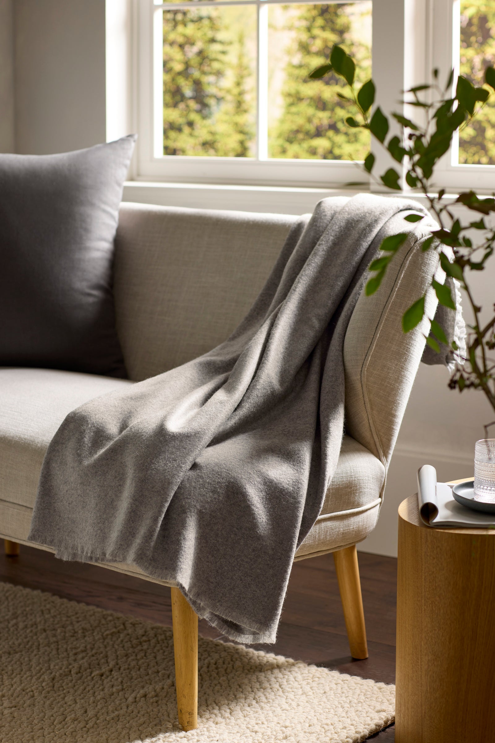 A cozy living room scene with a light beige sofa, a Cozy Earth Cashmere Fringe Blanket in grey draped over the arm, a matching grey cushion, a wooden side table with a glass of water, and sunlight streaming through the window.
