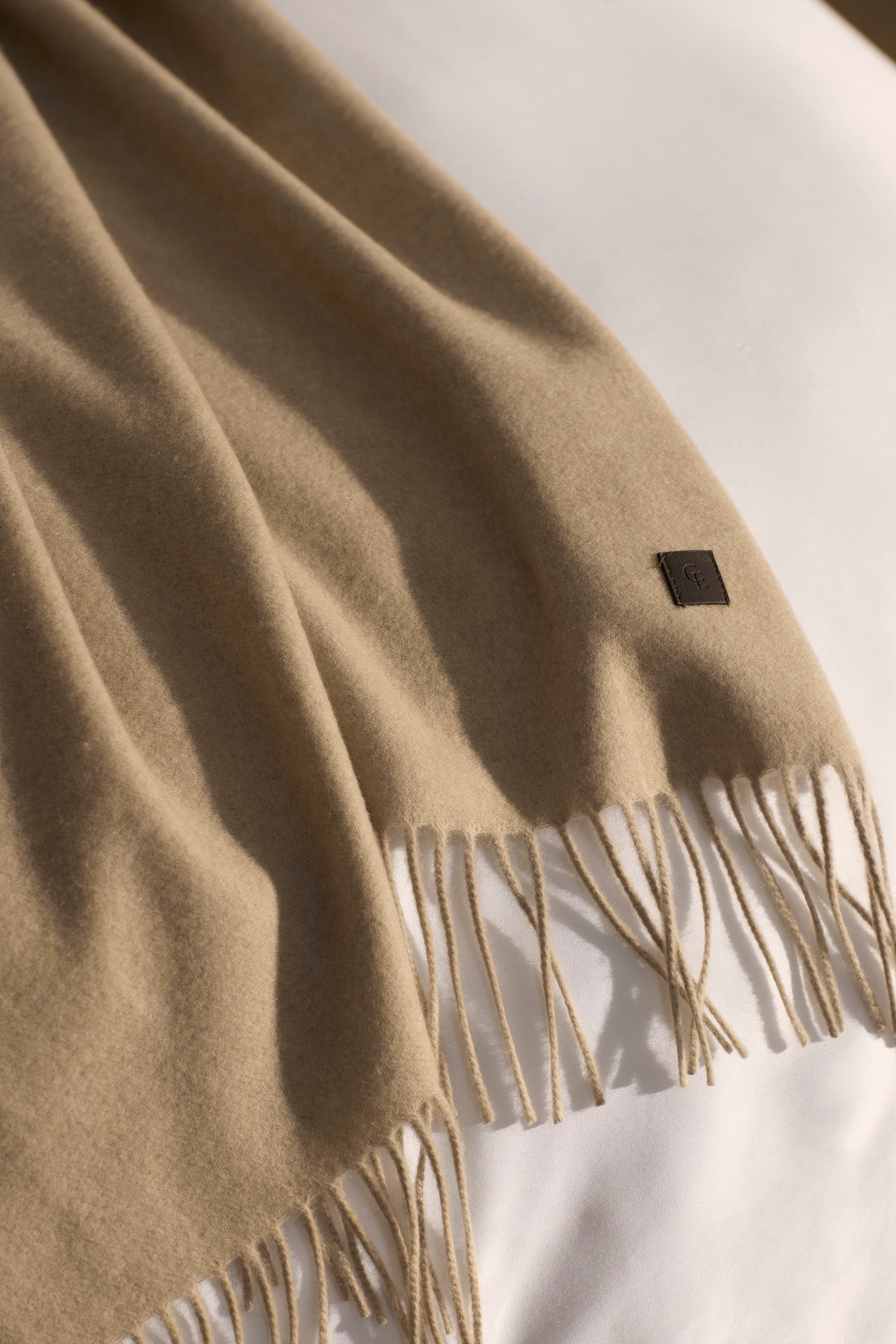 A soft, light brown Cashmere Tassel Throw by Cozy Earth is draped over a white surface. Near the fringes, a small dark rectangular tag with the logo is visible.|Color:Dune
