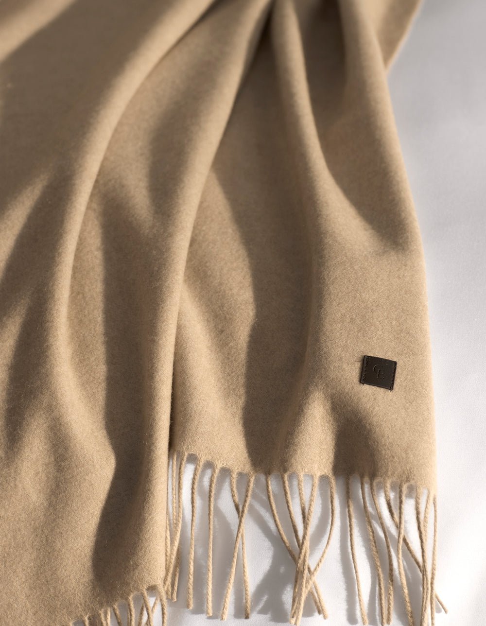 The Cashmere Tassel Throw by Cozy Earth, in soft beige with fringed ends and a small leather tag, is laid on a light surface as sunlight casts gentle shadows over its luxurious fabric.|Color:Dune