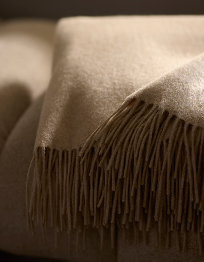 A close-up of the Cozy Earth Cashmere Tassel Throw, a soft beige blanket with tassel edges, neatly folded atop another in a warmly lit setting.