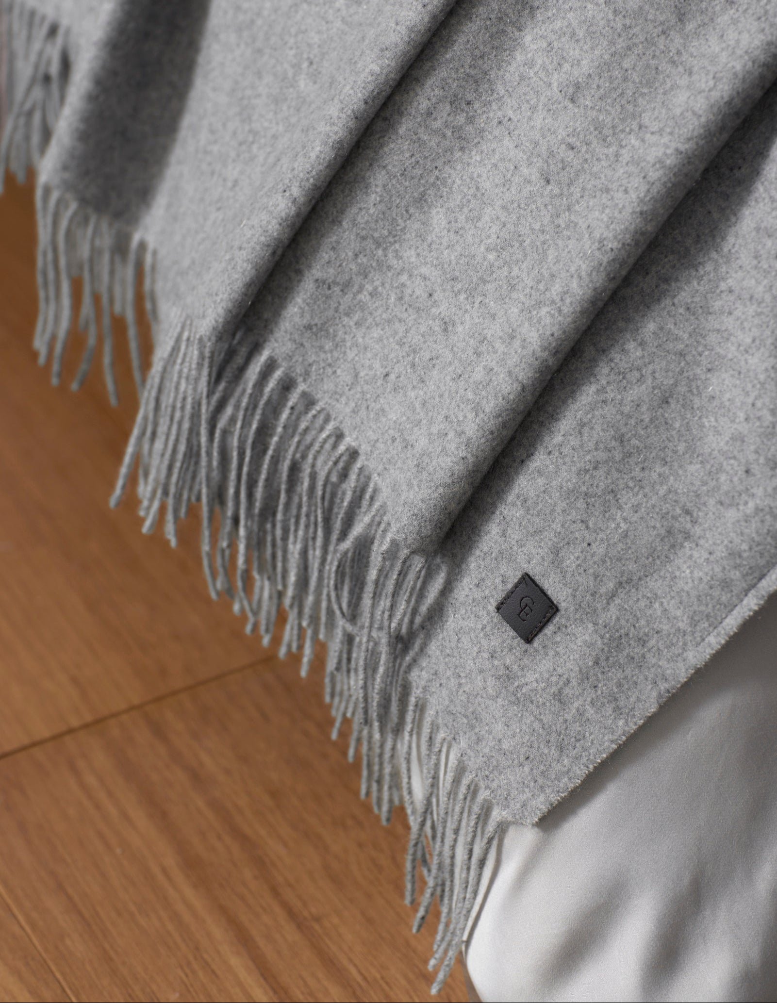 The Cozy Earth Cashmere Tassel Throw, a soft light gray blanket with fringed edges and a small black label, drapes over a bed edged in white sheets above a wooden floor.