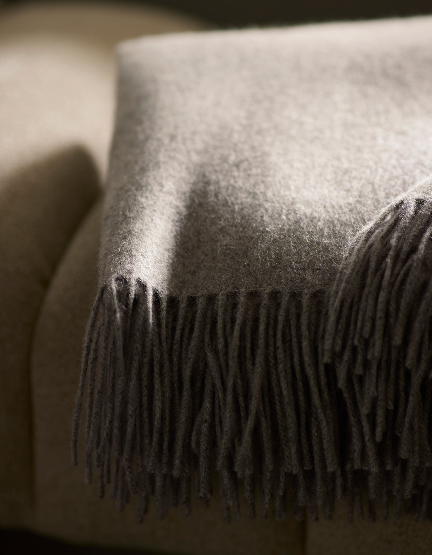 A close-up of the Cozy Earth Cashmere Tassel Throw, a soft gray blanket with fringed edges, draped over a beige upholstered sofa. Sunlight highlights its luxurious texture and fibers.