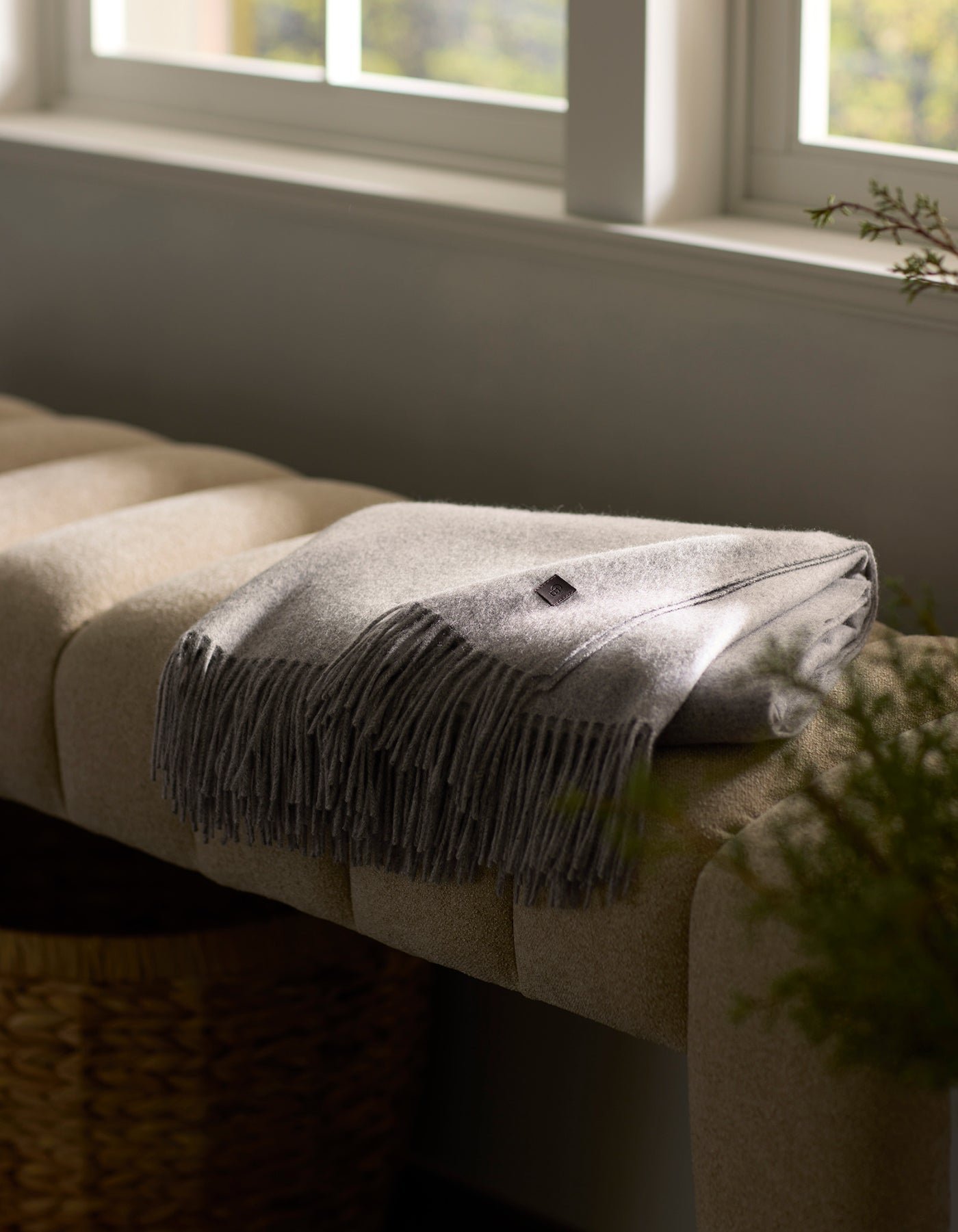 The Cozy Earth Cashmere Tassel Throw, in soft light gray, is neatly folded on a beige cushioned bench by a window. A woven basket peeks out below as warm sunlight fills the cozy room.