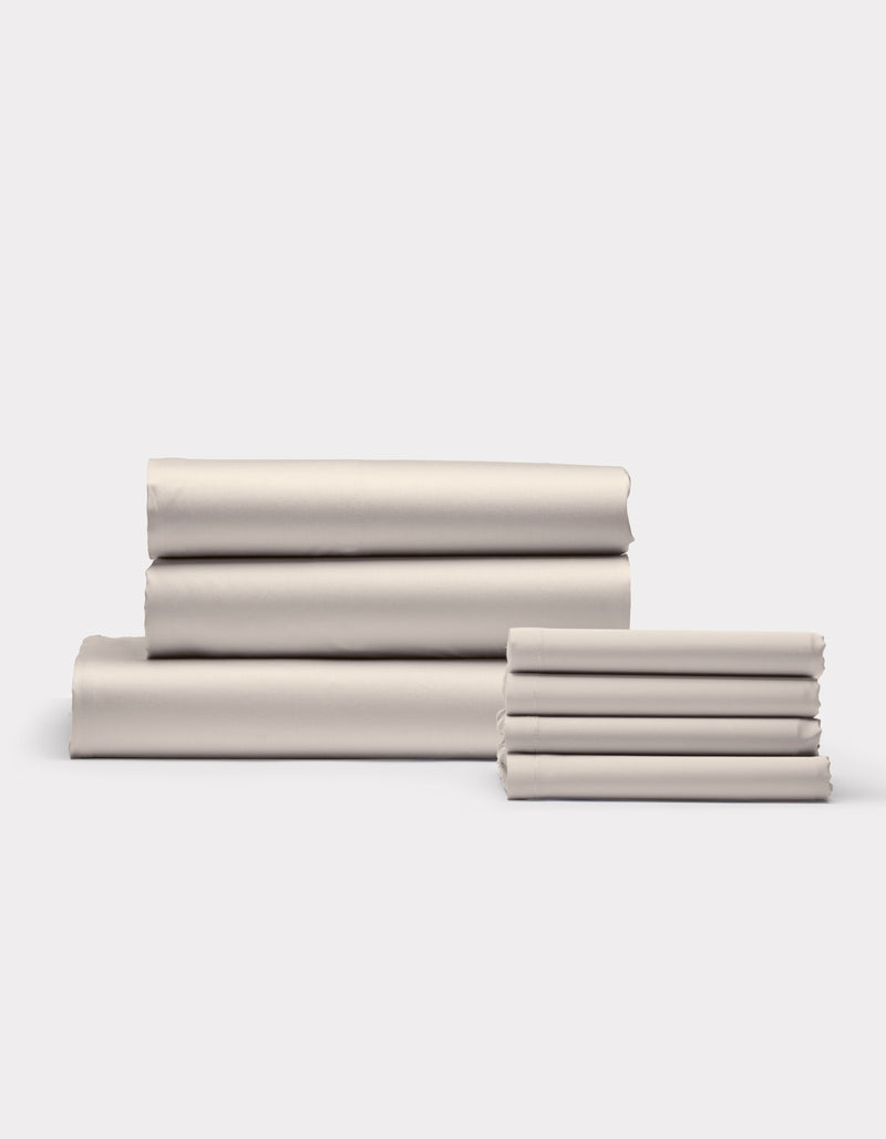 Cozy Earth's Bamboo Bedding Core Bundle features neatly folded, light beige bed sheets stacked in two piles on a plain white background.