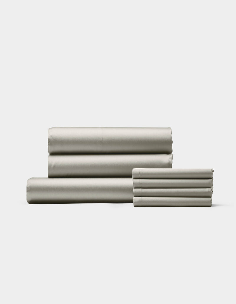 The Cozy Earth Bamboo Bedding Core Bundle, featuring neatly folded light gray sheets and pillowcases, is stacked on a white background with three larger sheets on the left and pillowcases on the right.