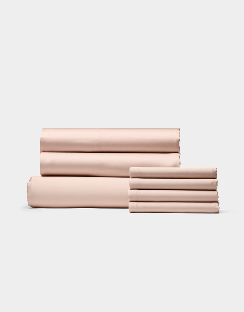 Cozy Earth’s Bamboo Bedding Core Bundle, shown in light pink, features neatly folded bed sheets and pillowcases arranged in two stacks against a simple, light background.
