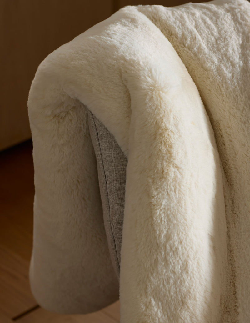 The Cozy Earth Cuddle Blanket, in soft cream-colored faux fur, is draped over a light gray chair, its cozy texture highlighted by natural light.