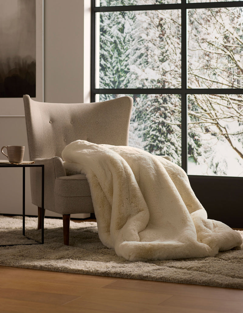A beige armchair with a Cozy Earth Cuddle Blanket is placed by a small table with a mug, set before large windows overlooking a snowy, tree-filled landscape.