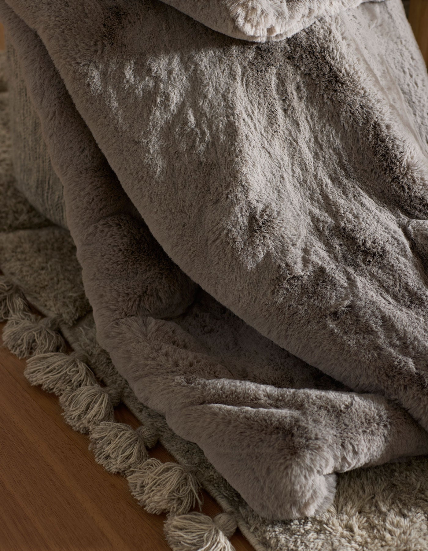 A close-up of the Cozy Earth Cuddle Blanket, a soft gray faux fur throw, draped over a textured beige rug with fringed edges on a wooden floor.