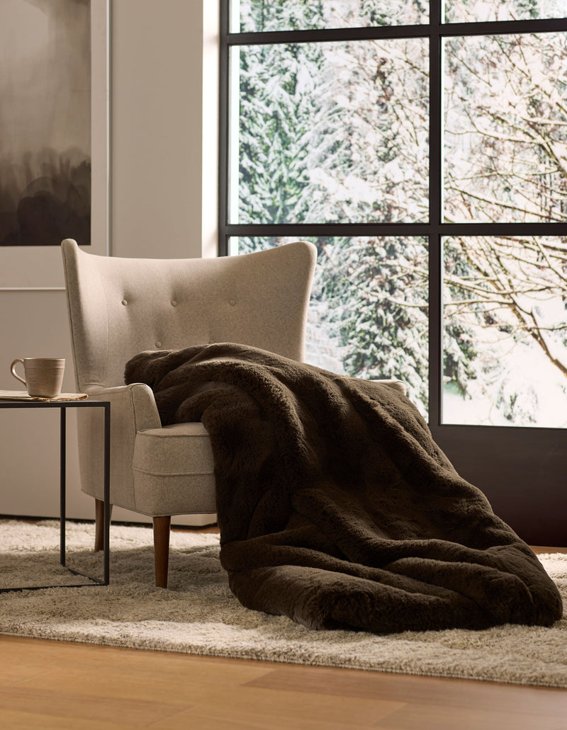A cozy beige armchair with a Cozy Earth Cuddle Blanket draped over it sits next to a small table with a mug, overlooking snowy trees through large windows.