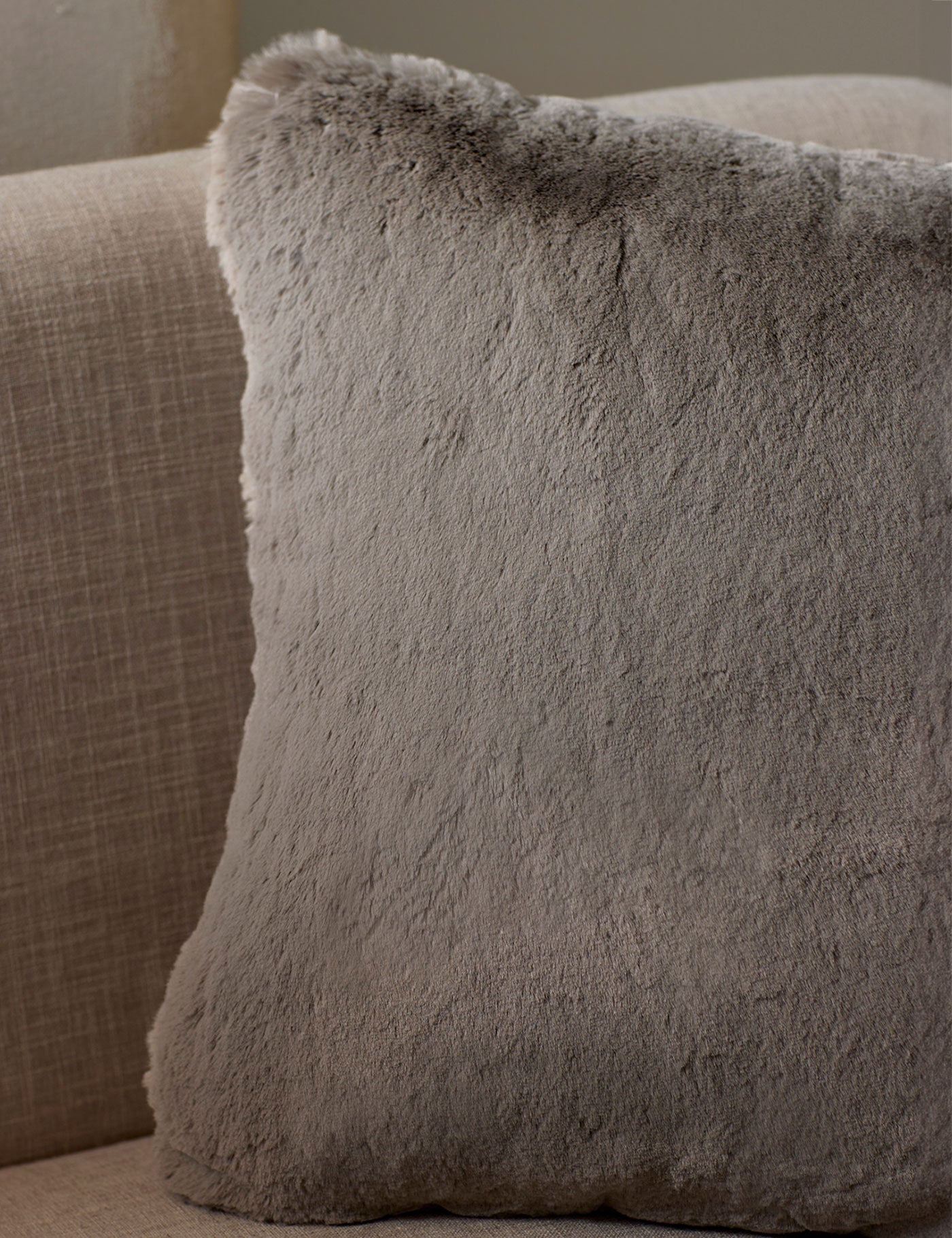 The Cozy Earth Cuddle Pillow, a large square in light gray faux fur, rests against the back of a beige fabric sofa.|Color:Dove Grey