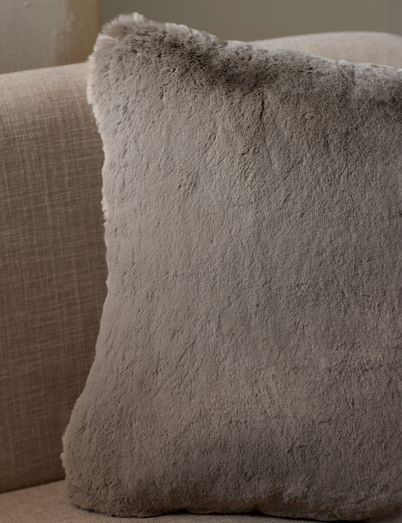 The Cozy Earth Cuddle Pillow, a large square in light gray faux fur, rests against the back of a beige fabric sofa.