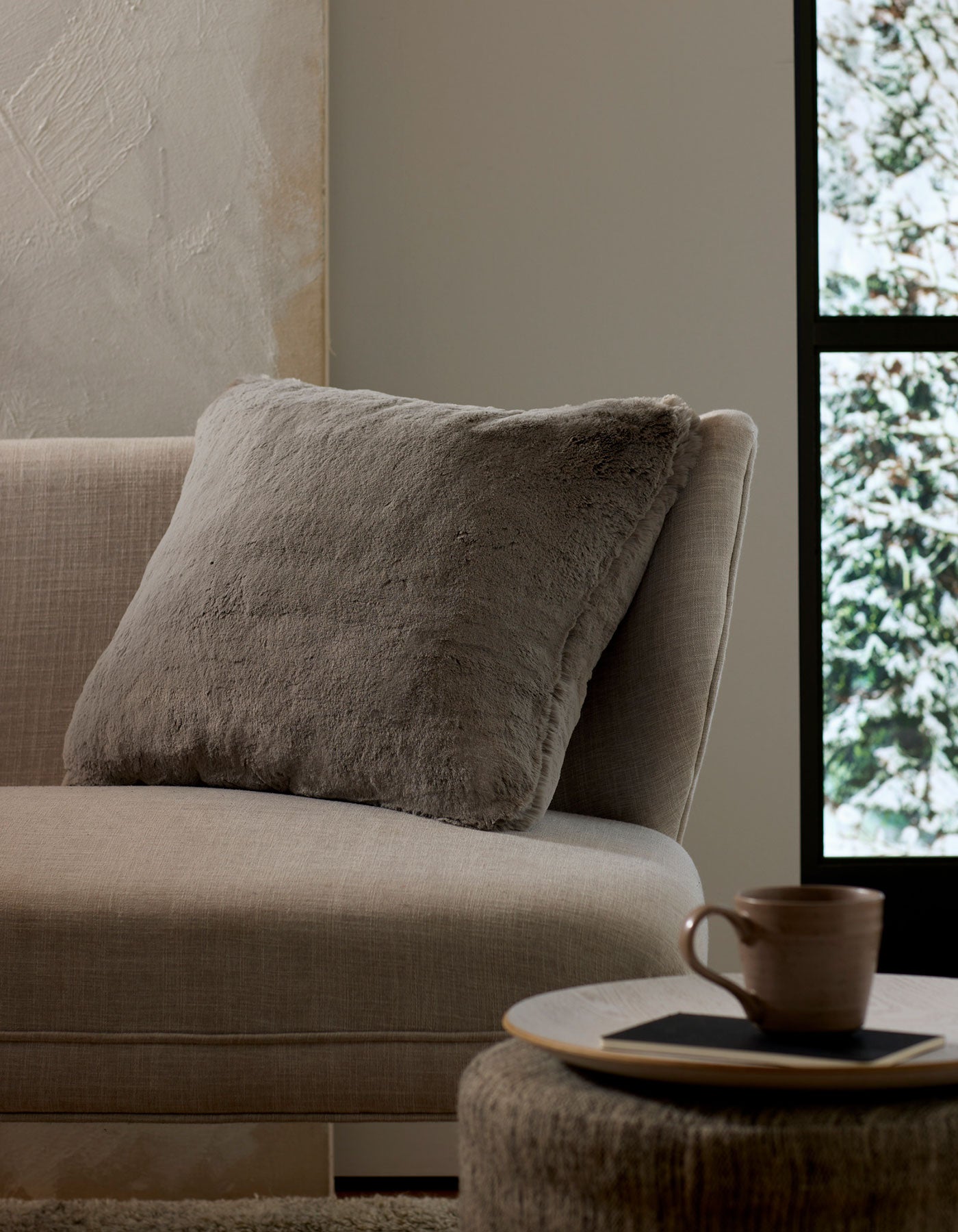A cozy beige couch with a large, soft Cuddle Pillow by Cozy Earth rests beside a round table holding a ceramic mug and coaster. Sunlight pours through the window, revealing snowy trees outside.