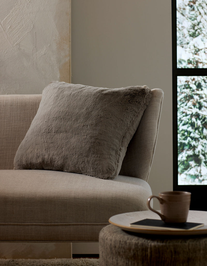 A cozy beige couch with a large, soft Cuddle Pillow by Cozy Earth rests beside a round table holding a ceramic mug and coaster. Sunlight pours through the window, revealing snowy trees outside.