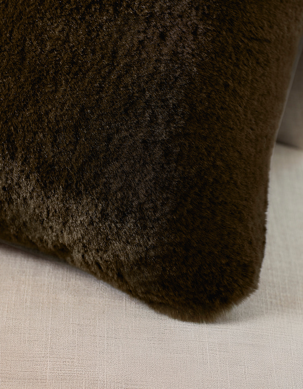 A Cozy Earth Cuddle Pillow in dark brown rests against the back of a light beige, textured fabric sofa.