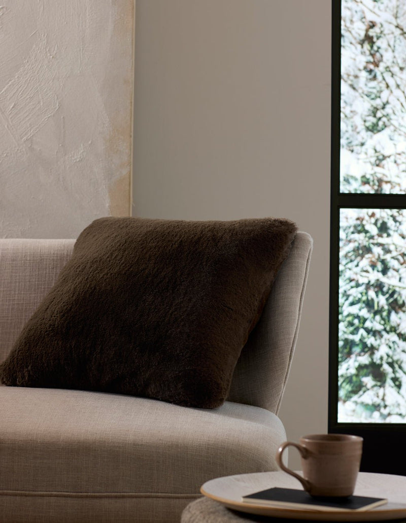 A beige sofa with the Cozy Earth Cuddle Pillow in dark brown sits by a window overlooking snow, while a ceramic cup and saucer rest on a small table nearby.