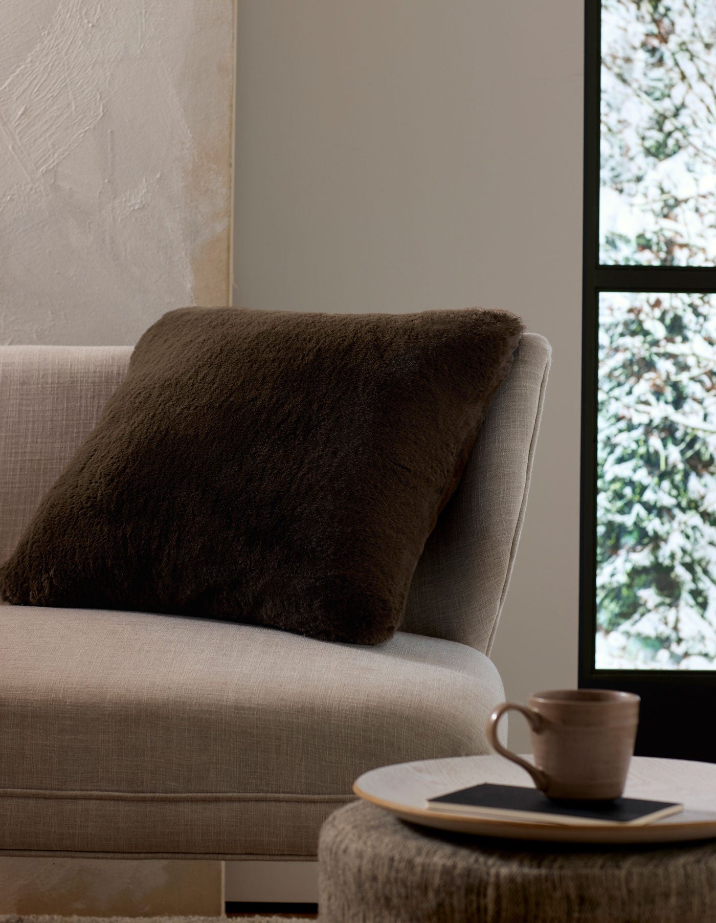 A cozy beige sofa with a Cozy Earth Cuddle Pillow in dark brown is beside a snowy window. In the foreground, a ceramic mug rests on a small round table with a saucer and black napkin.
