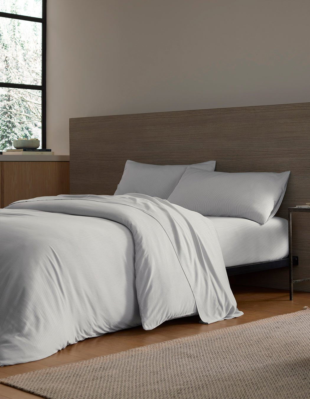 A modern, minimal bedroom features a neatly made bed with Cozy Earth’s Bamboo Bedding Deluxe Bundle against a wooden headboard. White bedding and pillows, a woven rug, and a small bedside table complete the cozy look.