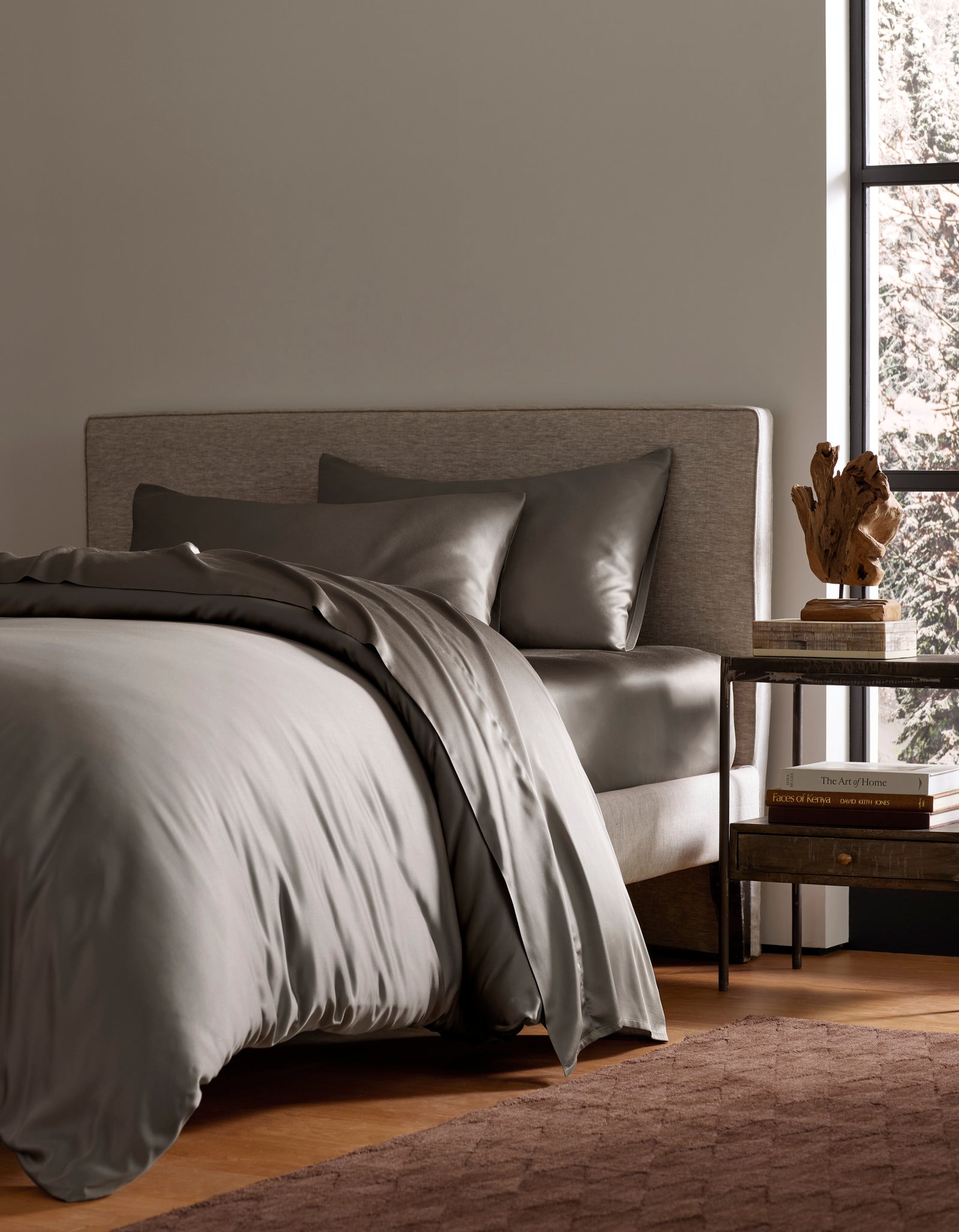 A neatly made bed features the Bamboo Bedding Deluxe Bundle by Cozy Earth in gray, two matching pillows, a light upholstered headboard, a wooden nightstand with books and a sculpture, and natural light streaming through a large window.