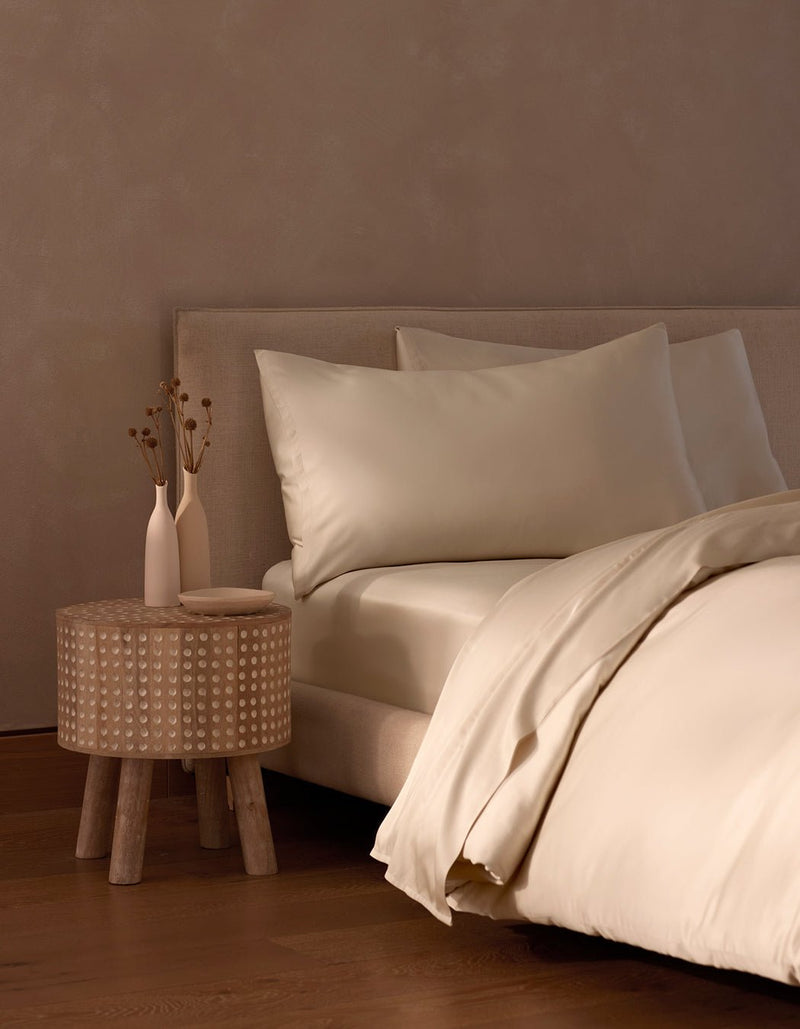 A neatly made bed with Cozy Earth’s Bamboo Bedding Core Bundle sits beside a small wooden nightstand holding two white vases with dried flowers. Cream-colored sheets and pillows complete the room’s minimalist, neutral-toned decor.