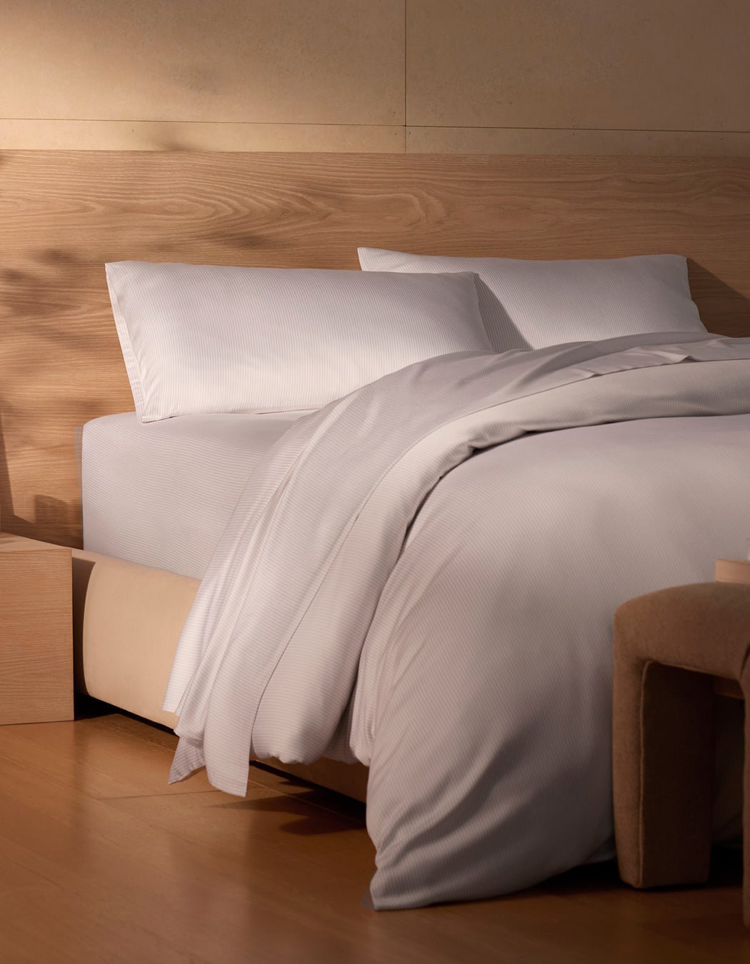 A neatly made bed features Cozy Earth's Bamboo Sheet Set in white, paired with pillows, a wooden headboard, light wood floors, and a beige upholstered bench in a softly lit minimalist bedroom.