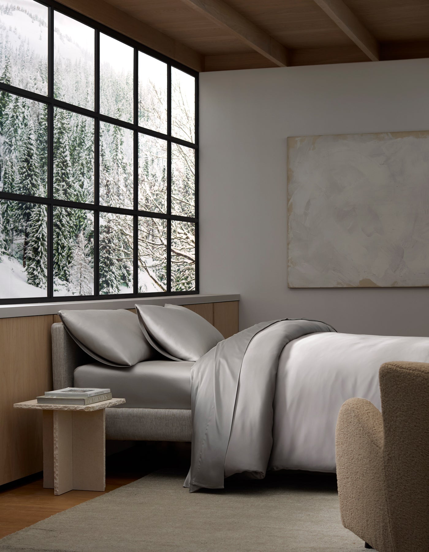 A cozy bedroom filled with natural light features the Cozy Earth Bamboo Bedding Deluxe Bundle on the bed, gray sheets and pillows, a small table, a beige chair, a large window overlooking a snowy forest, and a blank canvas on the wall.