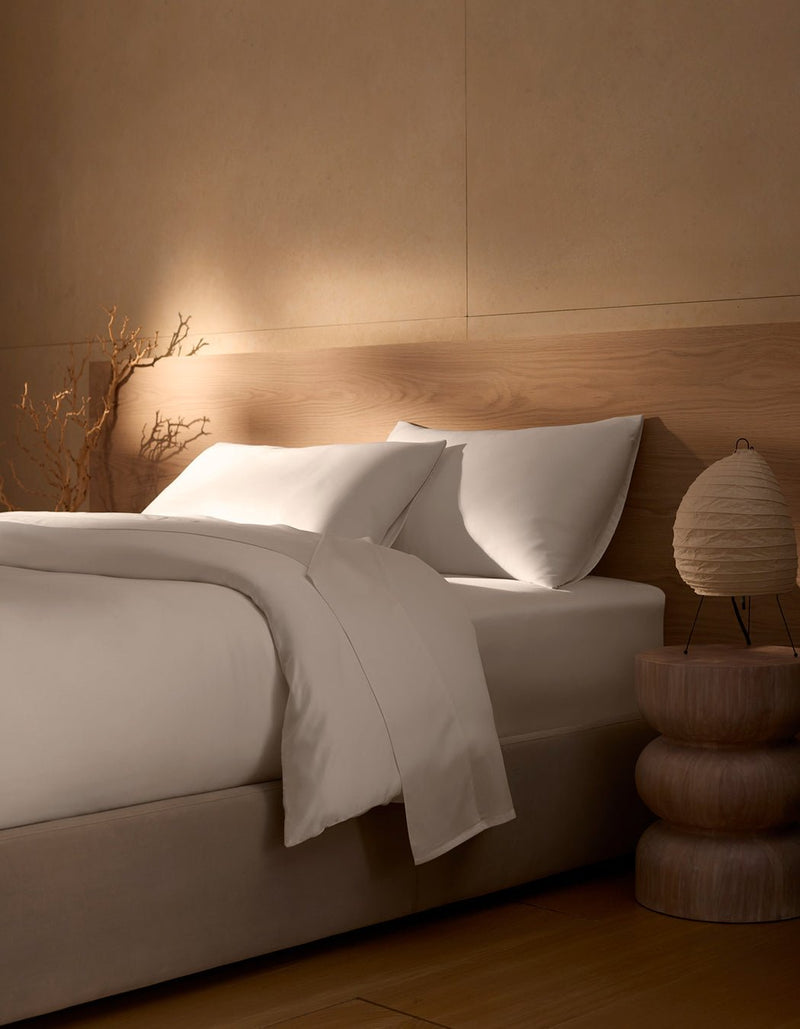 A neatly made bed featuring Cozy Earth's Bamboo Duvet Cover is set beside a wooden nightstand with a round lamp; beige walls, natural wood accents, and a dried branch in the corner create a calm, minimalist atmosphere.